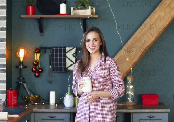 Happy young woman in plaid pajama holding jar smiling at camera in kitchen decorated for Xmas