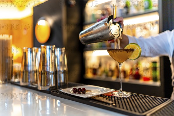 Hand of a waiter pouring cocktail into martini glass garnished with lemon in the counter of a