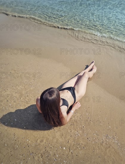 Charming young woman in bikini lying and tanning on the beach with legs stretched to the sea. Clear
