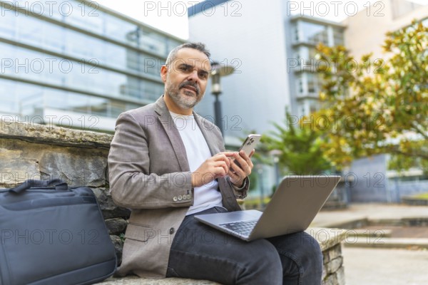 Mature male digital nomad working with laptop and mobile phone outdoors in the city