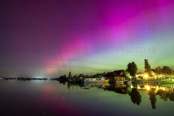 Calm lake with vivid auroras reflected next to illuminated buildings, harbour, Allensbach, Lake