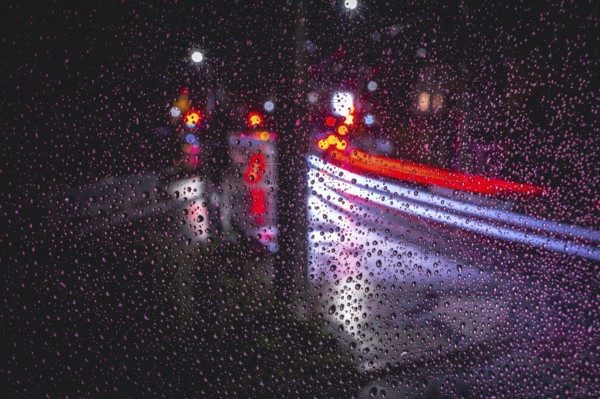 Rain falls on a pane of glass, blurred traces of light from vehicles and street lamps at night