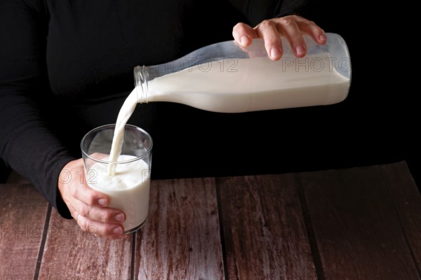 Hands of a woman pouring fresh milk into a glass, with a glass bottle