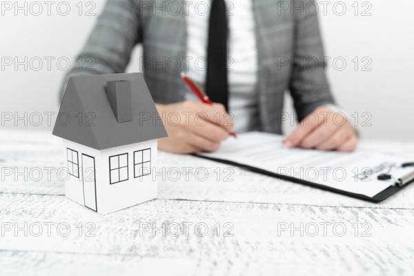 Businesswoman sitting and writing in notebook. Paper house on desk