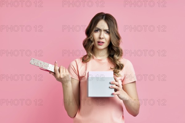 Young woman disappointed with her gift box on pink background. Bad present, emotion concept