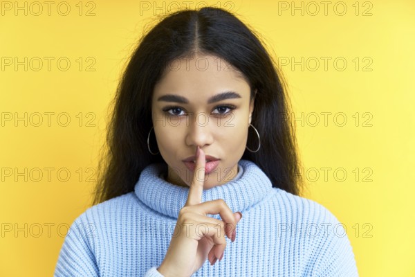 Pretty black woman keeps finger on lips, making hush gesture on yellow background. Shh, silence