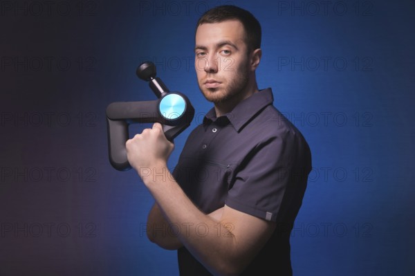 Portrait of a confident professional masseur with a percussion massager in his hands. Low key, blue