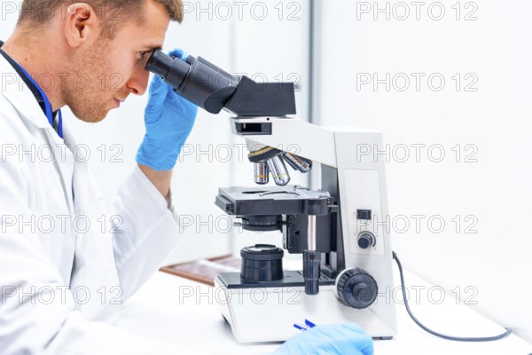 Side close-up view of a male young doctor using microscope to analyze samples in a laboratory
