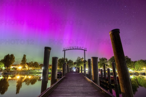 A wooden walkway under a glowing sky with colourful lights and trees with auroras, harbour,