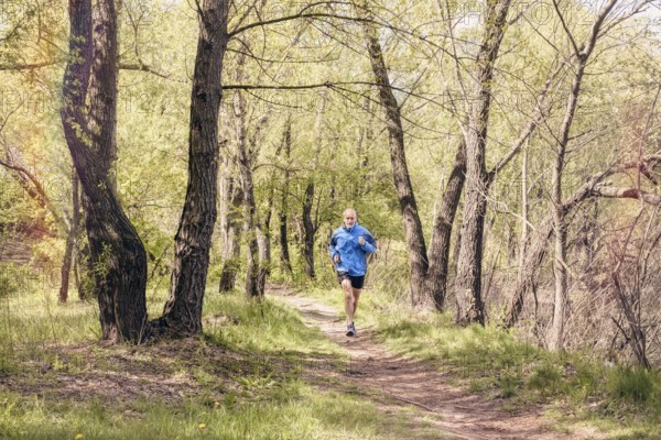 A senior man worn in black and blue is running in the forest, during a warm spring day