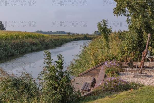 A quiet river flows along a bank with benches, flowers and trees, krummhörn, east frisia, germany
