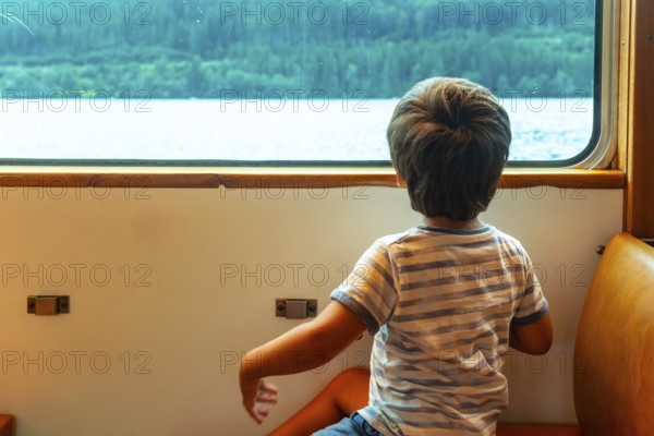 A young boy is sitting on a train window looking out at the water. The boy is wearing a striped
