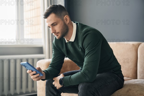 Relaxed man sitting on couch using hsi mobile smart phone at home. Social media concept
