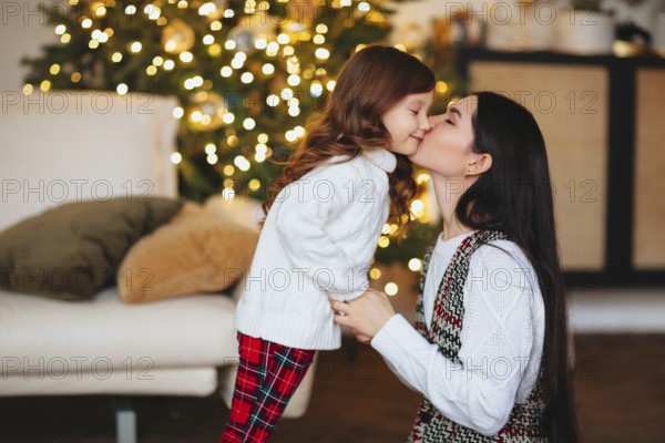 Beautiful loving mother gently kissing cute little daughter against festively decorated Christmas