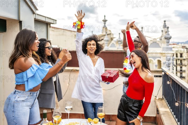 Happy man receiving a birthday gift celebrating with multi-ethnic friends during sunset rooftop