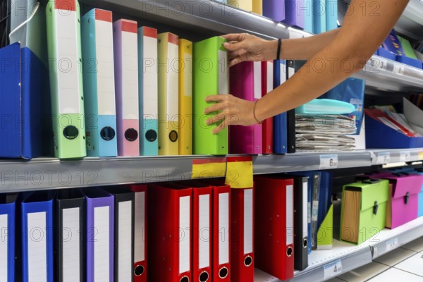 A person is reaching for a green binder on a shelf. The shelf is full of colorful binders and