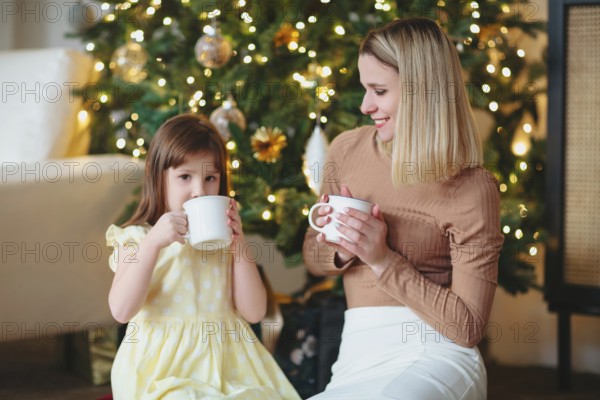 Happy family, mother and little daughter drinking hot cacao, sitting near decorated ?hristmas tree,