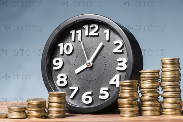 Round alarm clock with stack increasing coins against blue background. High resolution photo