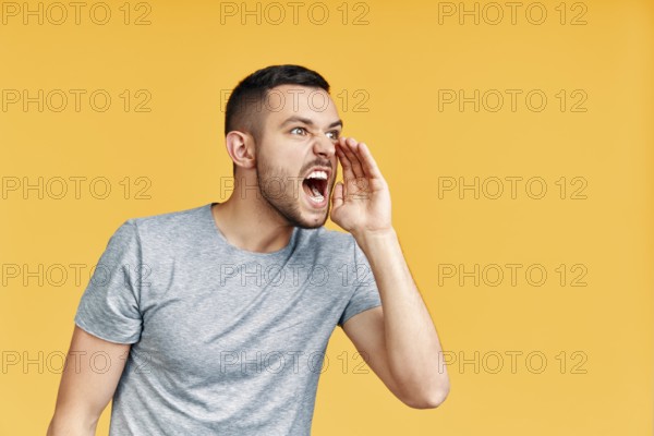 Young angry man shouting and screaming loud to side with hand on mouth and copy space for text over