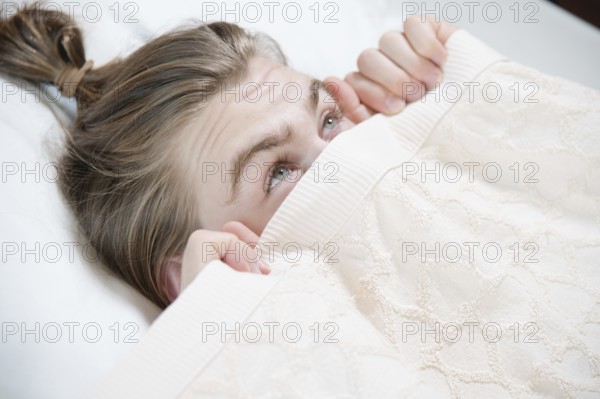 A young attractive man peeks out from under the blankets hiding behind them with fear when he wakes