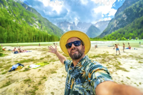 Happy man in a hawaiian shirt and straw hat taking a selfie at lago di landro, with tourists and