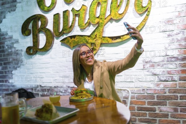 Happy latin blonde woman taking selfie while eating a tasty hamburger at fast food restaurant