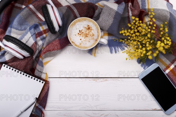Hot coffee cappucino, blanket, mimosa flowers, notepad, smartphone and head phones on old wooden