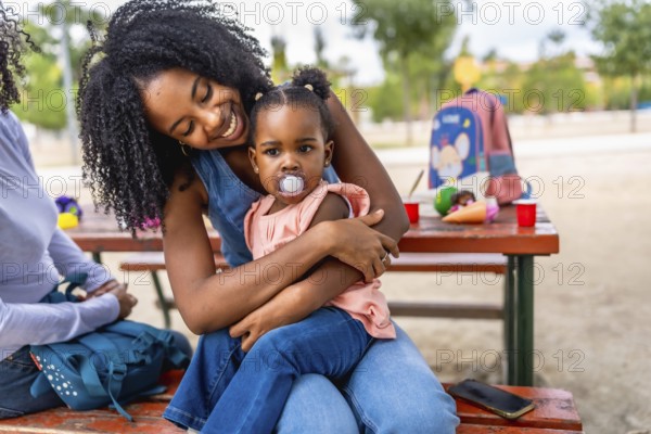 Happy african mother sitting on picnic table embracing a baby girl with pacifier in the park
