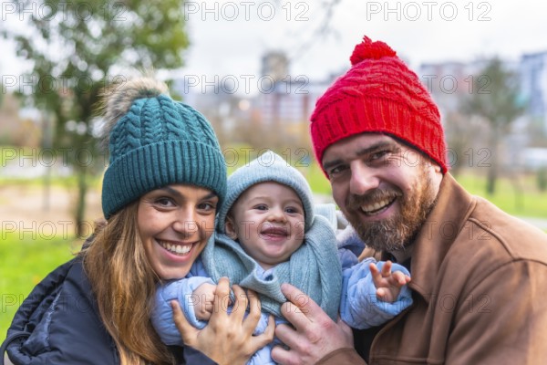 Smiling parents holding their baby wearing winter clothes and wool hats, enjoying a sunny winter