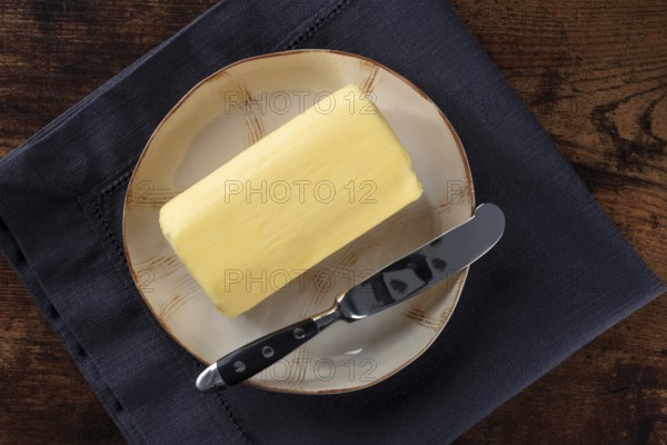 A block of butter with a knife on a plate, overhead flat lay shot on a dark rustic wooden table