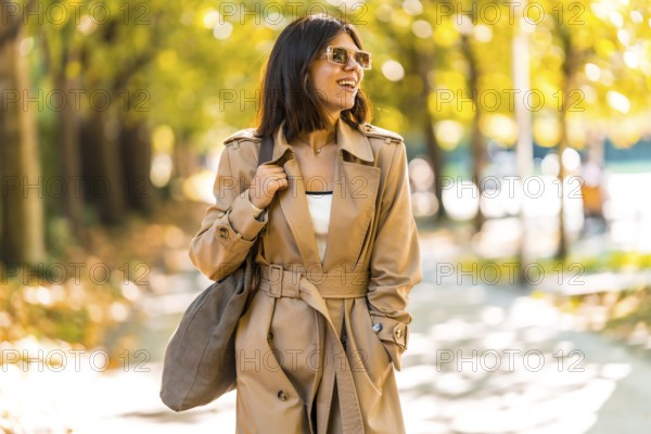 A woman wearing a tan trench coat and sunglasses walks down a sidewalk. She is smiling and carrying
