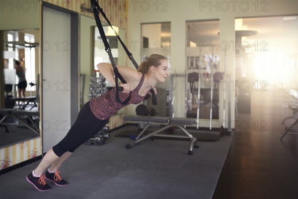 Young fit blond woman exercising with suspension bands in a modern gym in the morning
