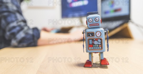 Toy robot on a desktop, man working on the computer in the background. Symbol for artificial