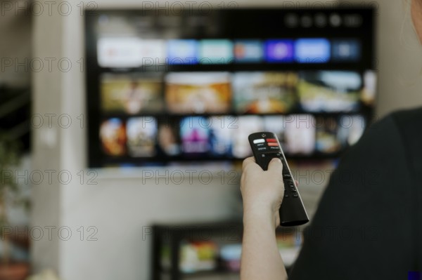 Hand holding remote control pointing to TV. Person watching smart television indoors. Blurred