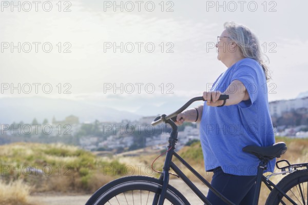 An overweight mature woman rides a black retro-style bicycle against a cloudy mountain backdrop