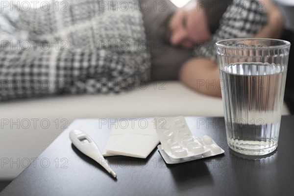 meds, medical thermometer and glass of water on nightstand against sick person suffering from flu
