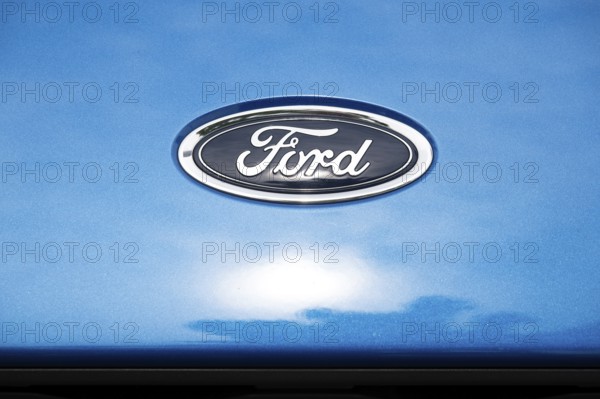 Logo of the car manufacturer Ford, sun behind clouds reflected in the paintwork