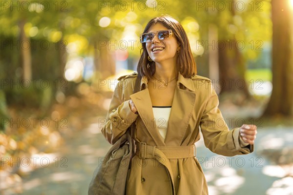 A woman wearing a tan trench coat and sunglasses is walking down a path. She is smiling and she is