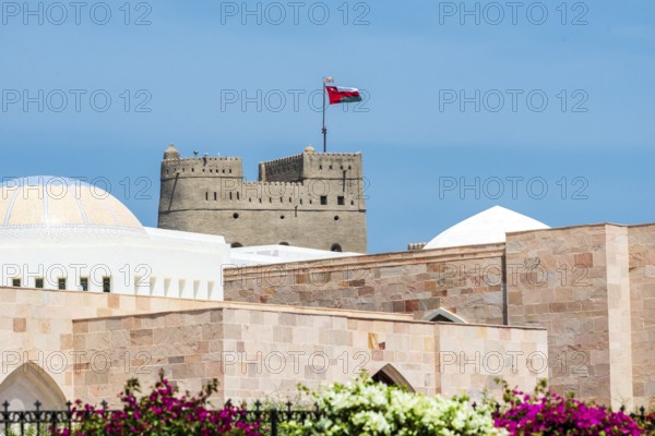 An old fortress with the Omani flag rises above pink stone buildings under a blue sky, Al Alam