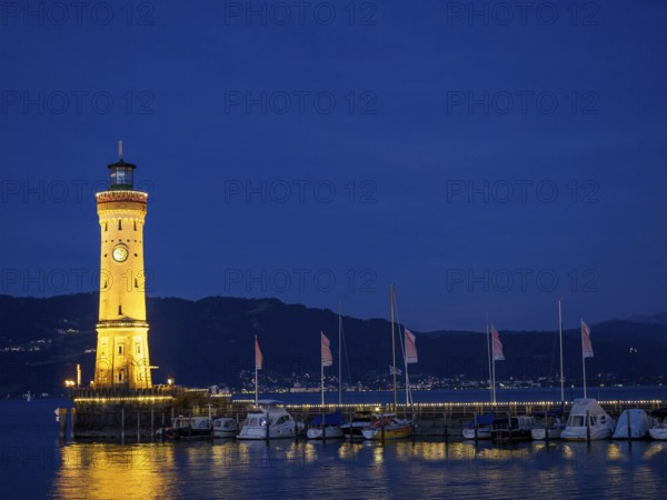 Illuminated lighthouse in the harbour at night, calm water and boats illuminated with warm light,