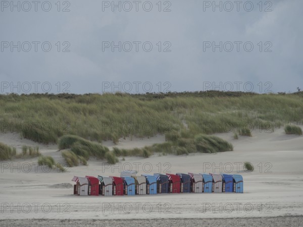 Row of colourful beach chairs along a sandy beach with high dunes in the background under a cloudy