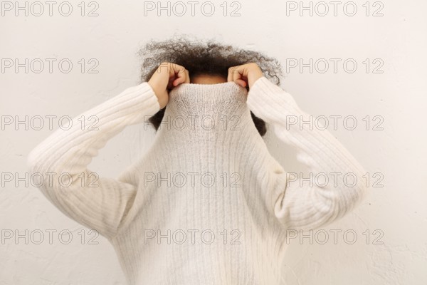 African curly female covering face with warm white sweater collar isolated on light studio