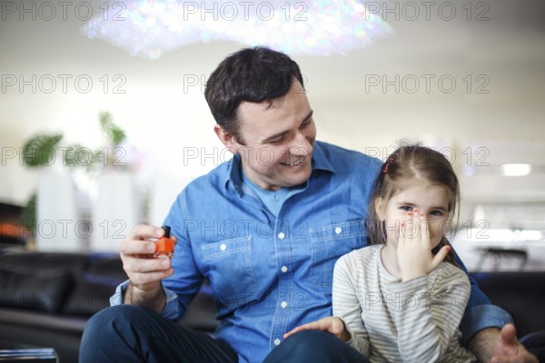 Cheerful father playing with little daughter sitting on couch and helping to paint her nails with