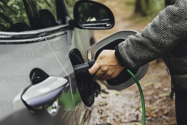 Hand connects an electric car to a charging plug, surrounded by a forest, environmentally friendly