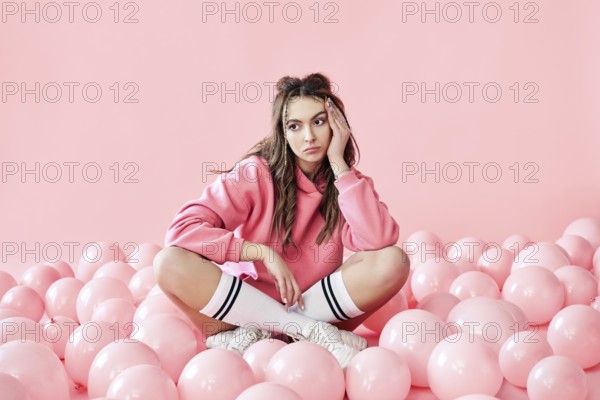 Bored fashionable woman sitting on floor with pink air balloons on pink pastel background.