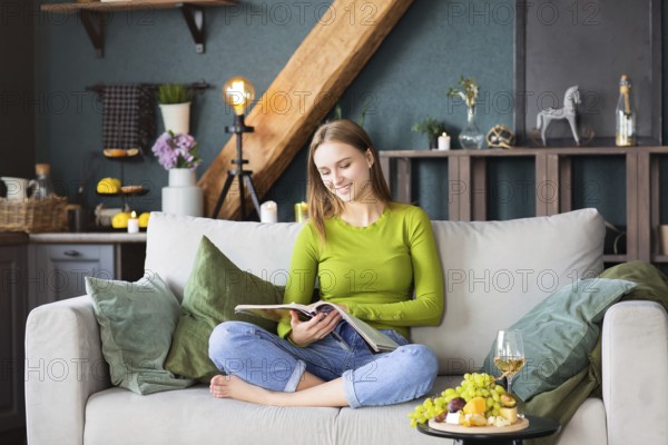 Young blond female reading interesting magazine while sitting on comfortable couch near table with