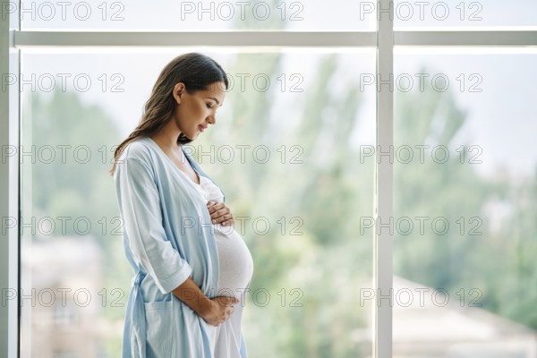 Portrait of young pregnant woman standing near window holding hands on her belly at modern home
