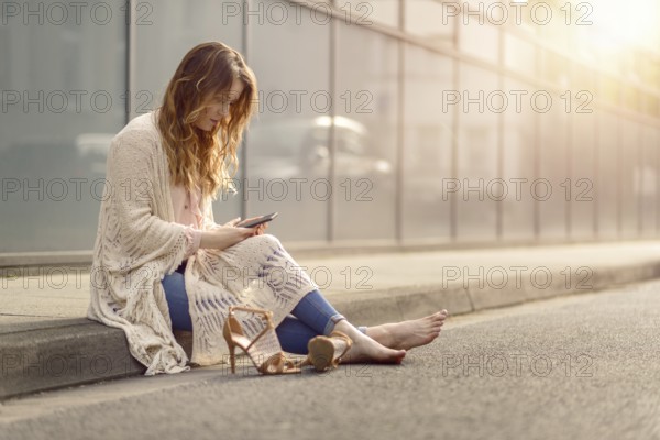 Attractive trendy woman sitting on the kerb of an urban street with her high heeled shoes lying on