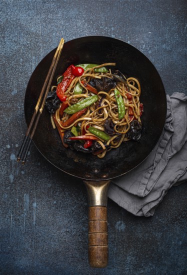 Asian dish stir fry udon noodles with vegetables and mushrooms in black rustic wok pan with wooden