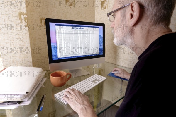 Mature man with beard working on a spreadsheet on computer. Folders with files, pens and an orange